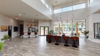 This contemporary single-floor community space is designed with an open, airy layout that immediately impresses upon entry. The welcoming reception lobby features large floor-to-ceiling windows, double glass-panel entry doors, and light wood-look flooring that flows seamlessly throughout, flooding the space with natural light and lending a clean, modern aesthetic. Tasteful beach-inspired décor, including coastal artwork, surf accents, and lush greenery, adds warmth and personality to the neutral palette.

At the heart of the space is a prominent reception and leasing counter finished in dark cabinetry with a granite surface, lined with swivel bar-height chairs and anchored beneath a dramatic constellation-style pendant chandelier. The double-height ceiling in this central area amplifies the sense of scale, with a clerestory window band drawing in additional light from above.

Adjacent to the main hall, a fully equipped kitchen with stainless steel appliances and additional pendant lighting serves as a functional amenity hub. Sliding glass doors open directly to an outdoor pool area, seamlessly connecting indoor and outdoor living. A separate lounge area features shiplap wall detailing, a large wall-mounted television, and comfortable seating, offering a relaxed retreat. Private offices or meeting rooms branch off a connecting corridor, completing this well-appointed community facility.