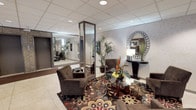 This single-floor lobby presents a thoughtfully designed common area that balances functionality with refined aesthetics. The space is anchored by a comfortable seating arrangement comprising dark brown upholstered armchairs grouped around a glass-topped coffee table adorned with floral arrangements, all set atop a bold, richly patterned area rug with red, gold, and black motifs against terrazzo flooring.

A distinctive mirrored column serves as a central architectural feature, visually expanding the space and adding depth. Complementing this, a decorative oval mirror with an ornate silver frame is mounted above a sleek console table along a textured accent wall, accompanied by tropical potted plants that introduce natural elements throughout.

The lobby also incorporates a dedicated mailroom area, housing a full bank of individual mail slots along one wall, with a bench and a glazed interior door providing access to an adjacent office or management space. A separate hallway leads to a secondary area featuring patterned wallcovering, framed artwork, a decorative stone planter, and additional mirrored paneling near what appears to be elevator access. Recessed lighting throughout ensures consistent, even illumination across the entire lobby.
