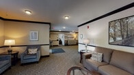 Located at 5183 Southpoint Dr in Louisville, KY, this single-floor facility presents a thoughtfully designed lobby and common area that balances comfort with a professional, welcoming atmosphere. The entry foyer features automatic sliding glass doors that flood the space with natural light and offer views of the surrounding greenery outside. A decorative wall clock and an information console greet visitors upon arrival, setting a refined tone from the moment of entry.

The open lobby extends into a generously proportioned lounge area carpeted throughout, with warm neutral tones and dark crown molding accenting the walls and ceiling. Seating areas are strategically arranged to encourage relaxation and conversation, featuring plush sofas, cushioned armchairs, and side tables with elegant table lamps. Framed artwork depicting serene outdoor scenes adorns the walls, contributing to the calm, inviting ambiance.

A standout feature of the lounge is a striking stacked-stone accent wall housing a fireplace and a wall-mounted flat-screen television, flanked by built-in shelving with decorative plants and accessories. This focal point anchors the room and adds a warm, residential quality to the space. Near the windows, a pair of upholstered accent chairs and a side table create a quiet nook bathed in natural light. Together, these elements compose a cohesive, comfortable environment suited for gathering and relaxation.