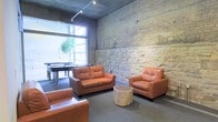 This single-floor space combines industrial architectural elements with comfortable, casual furnishings to create a welcoming lounge environment. The most striking feature is the expansive exposed stone wall, which spans the length of the room and adds raw, textural character to the space. Complementing this, a concrete ceiling with track lighting provides both ambient and focused illumination throughout the area.

The lounge area is anchored by a seating arrangement of tan leather sofas and armchairs, arranged around a natural wood stump coffee table, lending an organic touch to the otherwise industrial aesthetic. The flooring is finished in a neutral grey carpet, adding warmth underfoot.

Toward the back of the space, large industrial-style windows allow abundant natural light to flow in, offering views of greenery outside and enhancing the open feel of the room. Adjacent to the windows, a pool table is visible, indicating a dedicated games area that adds a recreational dimension to the space.

Overall, the combination of historic stone, modern track lighting, leather furnishings, and recreational amenities gives this space a dynamic, multi-purpose character suited for socializing and leisure.
