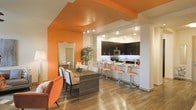 This single-floor space makes an immediate impression with its bold, energetic design aesthetic. The open-concept layout seamlessly connects the dining area and kitchen, creating a cohesive and functional living environment. A striking orange accent wall and matching ceiling detail serve as the visual centerpiece of the space, injecting personality and warmth into the interior.

The dining area is well-appointed with a substantial rectangular dining table surrounded by a mix of upholstered and slatted chairs, offering both style and comfort. An orange armchair adds a playful, coordinated accent near the entrance, complementing the overall color scheme.

The kitchen is positioned at the rear in a pass-through configuration, defined by dark espresso cabinetry that contrasts sharply with the white walls and light countertops. Stainless steel appliances, including a microwave and range, add a modern, polished touch. Under-cabinet lighting lends a refined ambiance to the cooking space. A raised breakfast bar with sleek orange upholstered bar stools bridges the kitchen and living areas, providing an additional casual seating option.

Rich wood-tone flooring flows throughout the entire space, grounding the vibrant color palette while adding warmth. The overall ambiance is modern, lively, and thoughtfully designed, balancing bold visual choices with practical, well-organized living spaces.