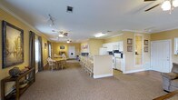 This single-floor clubhouse offers a bright and functional communal space designed for gatherings and events. The open-concept layout seamlessly connects the kitchen and dining areas, creating an inviting environment for residents and guests alike.

The kitchen is well-equipped with white cabinetry, a full-size refrigerator, a range, and a microwave, providing everything needed to prepare and serve meals for a group. A peninsula counter with seating extends into the main area, offering additional casual dining space and a natural transition between the kitchen and the rest of the room.

The dining and gathering area is generously sized, accommodating multiple tables and sets of wooden chairs, making it well-suited for group meals or community events. Warm yellow walls lend a cheerful and welcoming ambiance throughout the space, complemented by crown molding that adds a touch of classic detail. Neutral carpet flooring runs throughout, contributing to a comfortable and cohesive feel.

Ceiling fans with light fixtures are positioned throughout the room to ensure comfortable airflow, while track lighting provides additional illumination. Framed artwork decorates the walls, adding a homey character to the space. A private room or office is accessible through a door at the far end, offering additional functionality for the clubhouse.