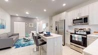 This two-story home offers a clean, contemporary aesthetic with a coastal flair throughout. The main floor presents an open-concept layout where the living area flows seamlessly into the kitchen and dining space. The kitchen is well-equipped with white shaker cabinetry, stainless steel appliances, a center island with bar seating, and recessed lighting, all set against large-format white tile flooring that extends throughout the main level. The living area is accented by ocean-inspired décor and a stylish area rug, creating a relaxed yet polished atmosphere.

The entry foyer welcomes guests with abundant natural light streaming through a full-lite door, complemented by coastal wall art and a decorative area rug. A staircase with carpeted treads connects the two floors.

The bedrooms are bright and well-proportioned, each featuring neutral walls, tile flooring, and windows that invite natural light. The primary bedroom includes an en-suite bathroom and a walk-in closet, while additional bedrooms share access to thoughtfully designed bathrooms. The bathrooms feature wood-tone vanities with white quartz countertops, large mirrors, glass-enclosed showers, and clean, modern fixtures. The overall ambiance throughout the home is light, airy, and cohesive, with a consistent coastal palette of whites, blues, and natural wood tones.