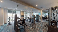 This fitness center is organized into several distinct zones, each thoughtfully designed to support a comprehensive workout experience. The main exercise floor is expansive, featuring patterned, multi-toned carpet in teal and earth tones that adds a dynamic visual element underfoot. This area is outfitted with a wide array of Matrix-brand strength machines, cable systems, free weights, and cardio equipment, including ellipticals and rowing machines. Large windows along one wall allow natural light to filter in through sheer curtains, creating an open, airy atmosphere, while floor-to-ceiling mirrors amplify the sense of space.

A separate strength zone houses a multi-rack system with weight plates and benches, positioned against mirrored walls and accessed through sleek black-framed glass doors. Adjacent to this is a dedicated cycling studio with wood-look flooring, a ceiling fan, bold teal-painted walls adorned with large-scale artwork, and a wall-mounted display screen for guided workouts. The studio's modern aesthetic is enhanced by recessed lighting throughout.

The entry foyer connects the zones via black-framed glass doors and features the same patterned carpeting, a built-in cabinetry station, and curated wall art, contributing to a cohesive, upscale aesthetic throughout the facility.