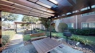 This thoughtfully designed outdoor community area showcases a range of amenities set within attractively landscaped grounds. The centerpiece is a resort-style swimming pool with a spacious brick-paved sundeck, furnished with blue lounge chairs and umbrella-shaded seating arrangements, ideal for relaxation. The pool area is framed by well-maintained greenery, mature trees, and manicured shrubs, all set against the backdrop of classic multi-story brick buildings featuring bay windows and private balconies.

A dedicated pergola structure provides a shaded gathering space, equipped with a charcoal grill, a wooden picnic table, and bench seating — making it well-suited for outdoor dining and social gatherings. The pergola's translucent paneled roof allows soft, diffused light to filter through while offering protection from the elements.

The broader courtyard features additional paved seating areas with café-style tables and chairs beneath market umbrellas, connected by well-kept walkways that wind through lush landscaping. Stone and mulched garden beds accent the grounds, adding texture and visual interest throughout. The overall ambiance is that of a cohesive, amenity-rich outdoor environment that balances comfort, functionality, and aesthetic appeal.