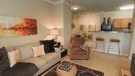 This single-floor residence offers a thoughtfully designed open-concept layout that connects the living, dining, and kitchen areas in a cohesive flow. The living area is anchored by neutral, warm-toned walls accented with crown molding, lending the space a refined, polished character. A gray sofa adorned with decorative pillows, including a distinctive coral motif cushion, pairs with a plush brown armchair to create a comfortable and stylish seating arrangement. A dark patterned area rug grounds the space, while a woven ottoman serves as a functional centerpiece.

Track lighting and a ceiling fixture provide well-distributed illumination, complementing the warm glow of a table lamp on the side table. Wall art in earthy red-orange tones adds a bold visual contrast to the muted backdrop.

Beyond the living area, a dining space is visible with a small table and chairs, and a mirror on the wall enhances the sense of openness. The kitchen features a raised bar-height counter with stylish stools, ideal for casual dining or entertaining. Overall, the space exudes a modern yet cozy ambiance, with consistent design elements tying each area together seamlessly.