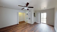 This 2-floor residence offers a clean, functional layout with thoughtful design details throughout. The main living area is highlighted by rich, dark hardwood flooring that adds warmth and character to the space. A ceiling fan with a classic wood-toned blade design is centrally positioned, providing both comfort and a finishing touch to the room. White walls keep the interior bright and neutral, allowing natural light to flow freely.

A sliding glass door fitted with vertical blinds opens to an outdoor area, filling the living space with additional natural light and providing convenient access to the exterior. The open sightlines connect the living area to adjacent rooms, including what appears to be a bedroom with a walk-in closet, offering generous storage. A glimpse into the kitchen area reveals additional functional space beyond the main living room.

Climate control is well-considered, with visible HVAC vents integrated into the walls. The overall ambiance is light, airy, and move-in ready, with a straightforward and practical layout that spans two floors, offering separation between living and private spaces. The combination of hardwood flooring, neutral tones, and natural light creates an inviting and comfortable environment throughout the home.