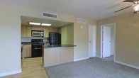 This single-floor residence offers a practical and open layout that seamlessly connects the kitchen and living areas. The kitchen is outfitted with light wood cabinetry, a dark granite-style countertop, and a full suite of black appliances including a range, microwave, and refrigerator. A peninsula counter extends into the space, providing an ideal spot for casual dining or meal prep while maintaining an open sightline into the living area. Recessed fluorescent lighting above the kitchen workspace ensures ample illumination. The living area features neutral carpet flooring, offering a warm contrast to the hardwood-style flooring in the kitchen. Walls are finished in a soft beige tone, contributing to the light and airy feel of the space. HVAC vents are positioned throughout the ceiling for efficient climate control. A hallway off the main living area leads to additional rooms, with white interior doors providing clean transitions between spaces. The overall design is straightforward and functional, making it a comfortable and easy-to-navigate living environment suited for everyday use.