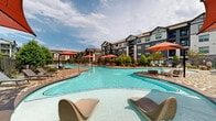 Located at 7303 S Peek Rd in Richmond, Texas, this outdoor amenity space showcases an impressive resort-style pool complex designed for both relaxation and recreation. The centerpiece is a series of generously sized, freeform swimming pools with crystal-clear blue water, connected across a sweeping, organically shaped layout that encourages leisurely movement throughout the space. The pool deck is finished in a classic herringbone-patterned brick, adding a refined, textured aesthetic underfoot that complements the surrounding architecture. Vibrant red market umbrellas are stationed throughout, providing shaded seating areas alongside rows of modern lounge chairs positioned to take full advantage of the open Texas sky. The space is framed on all sides by multi-story residential buildings featuring a craftsman-inspired exterior of stone, siding, and dark trim, creating an intimate, courtyard-like atmosphere. Mature trees, manicured shrubs, and potted plantings are thoughtfully integrated throughout the deck perimeter, softening the hardscape and enhancing the resort ambiance. Iron fencing defines the boundary of the pool area, offering both security and visual continuity with the surrounding green landscaping. The overall design creates a bright, welcoming outdoor retreat ideal for warm-weather enjoyment.