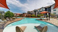 Located at 7303 S Peek Rd in Richmond, Texas, this outdoor amenity space showcases an impressive resort-style pool complex designed for both relaxation and recreation. The centerpiece is a series of generously sized, freeform swimming pools with crystal-clear blue water, connected across a sweeping, organically shaped layout that encourages leisurely movement throughout the space. The pool deck is finished in a classic herringbone-patterned brick, adding a refined, textured aesthetic underfoot that complements the surrounding architecture. Vibrant red market umbrellas are stationed throughout, providing shaded seating areas alongside rows of modern lounge chairs positioned to take full advantage of the open Texas sky. The space is framed on all sides by multi-story residential buildings featuring a craftsman-inspired exterior of stone, siding, and dark trim, creating an intimate, courtyard-like atmosphere. Mature trees, manicured shrubs, and potted plantings are thoughtfully integrated throughout the deck perimeter, softening the hardscape and enhancing the resort ambiance. Iron fencing defines the boundary of the pool area, offering both security and visual continuity with the surrounding green landscaping. The overall design creates a bright, welcoming outdoor retreat ideal for warm-weather enjoyment.