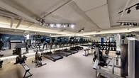 Located at 600 S Curson Ave, this fitness center offers a comprehensive workout facility within a single, open floor plan. The space is thoughtfully organized into distinct functional zones, making efficient use of its footprint. An exposed ceiling with visible ductwork and pipework lends an industrial aesthetic, while track lighting mounted on rails provides bright, focused illumination throughout.

The cardio zone features a row of TRUE-brand treadmills and stationary bikes, each equipped with individual display screens for an engaging workout experience. Adjacent to this area, a well-stocked free weights section houses multiple tiers of dumbbells on sturdy racks, accompanied by flat and adjustable benches for a variety of exercises. Full-length mirrors line the walls, enhancing the sense of space and allowing users to monitor their form.

A dedicated strength and functional training zone includes cable machines and resistance equipment, while a boxing corner features punching bags, medicine balls, and a wall-mounted gear rack. A bold cityscape mural adds a dynamic visual element to this area. The durable, speckled flooring throughout provides stability and ease of maintenance. The overall ambiance is clean, energetic, and purpose-built for a well-rounded fitness experience.