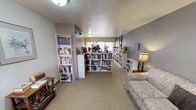 Located at 2215 Ambassador Rd NE in Albuquerque, New Mexico, this single-floor space functions as a well-organized lending library or community book exchange. The interior is finished with neutral gray walls, textured ceilings, and wall-to-wall carpeting throughout, creating a warm and comfortable environment for browsing and reading.

The main area is lined with an extensive collection of white bookshelves filled with a wide variety of titles, ranging from fiction and mysteries to general interest reads. The shelving units are thoughtfully arranged to define different zones within the open layout, maximizing the use of available floor space while keeping the area navigable and inviting.

Seating is a notable feature of this space. A patterned sofa anchors one section of the main room, while a separate nook offers two plush, dark upholstered wingback chairs arranged around a small side table with a floor lamp — ideal for quiet reading. Additional individual seating and lamps are positioned throughout, enhancing the cozy, reader-friendly ambiance.

Natural light filters in through curtained windows, complemented by flush-mount ceiling fixtures and table lamps that provide warm, even illumination. A door marked "Roof Access" is also present. The overall atmosphere is calm, organized, and welcoming, making this space a functional and charming destination for book lovers.