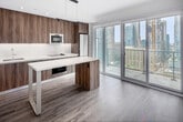 Located on the 24th floor at 8 Gloucester Street in the heart of Toronto's Church-Wellesley Village, this compact yet efficiently designed one-bedroom, one-bathroom unit offers a thoughtful modern layout across a single level. The open-concept living and kitchen area is anchored by dramatic floor-to-ceiling sliding glass doors that flood the space with natural light and frame sweeping views of the downtown Toronto skyline. Light wood-tone flooring runs throughout the main living areas, complementing the bright white walls and clean contemporary finishes. The kitchen is a standout feature, showcasing rich wood-grain cabinetry, integrated appliances including a built-in microwave and stainless steel refrigerator, a sleek cooktop, and a striking white waterfall-edge island with pendant lighting above — ideal for both meal preparation and casual dining. Track lighting in the living area adds a stylish, functional touch. The bedroom is a quiet, well-proportioned retreat with recessed lighting and the same warm hardwood flooring. The bathroom is finished with large-format beige floor tiles, full grey stone-effect wall tiling surrounding a soaking tub with an overhead rain shower, and clean white fixtures. A hallway connects the spaces efficiently, completing this well-appointed urban residence.
