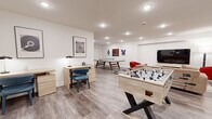 Located at 227 North Walnut in East Orange, NJ, this amenity lounge is a thoughtfully designed multipurpose space that balances recreation and productivity. The room features light wood-style flooring, crisp white walls, and recessed ceiling lighting that creates a bright, welcoming atmosphere throughout.

The space is divided into distinct functional zones. A lounge area anchors one side of the room, furnished with a neutral-toned sofa, bold red accent chairs, a wood coffee table, and a wall-mounted flat-screen TV set atop a light wood media console — ideal for relaxing or casual gatherings. Adjacent to the lounge, a foosball table and ping pong table provide ample entertainment options for residents.

A dedicated work zone features two light wood desks paired with blue leather chairs, each desk lamp-lit and accented by framed abstract artwork above — offering a quiet, focused environment for remote work or study.

The space is served by a wall-mounted mini-split HVAC unit, ensuring year-round comfort. A glass-paneled entry door with an illuminated exit sign provides easy building access. Decorative wall art is displayed throughout, adding visual interest and a curated, modern aesthetic. The open layout and cohesive design make this lounge a versatile and inviting community space.