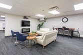Located at 3189 King St E in Kitchener, Ontario, this single-floor amenity space offers a thoughtfully designed recreational and social hub. The layout is divided into distinct zones that cater to both active recreation and relaxed gathering. The games area is anchored by a full-size pool table with a wall-mounted cue rack, set on stylish striped carpet tiles beneath a drop ceiling with recessed panel lighting that keeps the space bright and welcoming. An adjacent alcove features a dartboard mounted against a textured white brick-style accent wall, adding a playful touch to the space. The lounge area is warmly furnished with a white leather sofa and navy blue accent chairs arranged around a round wood coffee table, centered on a wall-mounted flat-screen television above a sleek linear electric fireplace — creating a comfortable focal point for socializing. A dining nook at the rear of the space includes a dark wood table with seating for several guests alongside a full-size stainless steel refrigerator, making it ideal for hosting. Throughout, the consistent grey carpet flooring, neutral white walls, and flush-panel ceiling lighting create a cohesive, modern aesthetic that balances comfort with functionality.