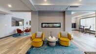 Located at 2 Broadway in Lynnfield, Massachusetts, this thoughtfully designed single-floor space offers an expansive open-concept layout with distinct functional zones that flow seamlessly into one another. The interior is anchored by soaring ceilings and wide-plank hardwood flooring throughout, lending the space an airy, welcoming atmosphere.

The living area showcases a curated modern aesthetic, featuring a wall-mounted television, a leather accent chair, and a colorful area rug that adds warmth and character. An interior pass-through window connects the living zone to adjacent areas, enhancing the open feel while maintaining visual interest. A bold gallery wall display of abstract artwork adds a creative focal point to the space.

At the heart of the layout sits a striking lounge area centered around a sleek, linear gas fireplace set into a full tile surround, flanked by vibrant mustard-yellow chairs — an inviting gathering spot that serves as a natural divider between zones.

The kitchen is outfitted with flat-front gray cabinetry, marble-look countertops, a stainless steel refrigerator, and a generous island with bar seating. Large windows with horizontal blinds flood the kitchen and adjacent dining nook with natural light and offer views of the surrounding landscape.

A secondary dining area with a counter-height table is positioned beside floor-to-ceiling windows, maximizing natural light and providing an open, connected feel throughout this well-appointed communal space.