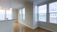This single-floor space is defined by clean architectural detailing and a bright, open atmosphere. The main living area features warm hardwood flooring that extends throughout, complemented by crisp white crown molding and tall baseboards that add a polished finish to the neutral, beige-toned walls. Multiple double-hung windows fitted with horizontal blinds line two walls, flooding the interior with generous natural light while offering views of surrounding greenery and the exterior streetscape. The windows are notably tall, reinforcing a sense of openness and airiness within the space. A partially visible countertop with granite-style surfacing suggests a kitchen or bar area adjacent to the main room, hinting at a practical and connected layout. Electrical outlets and switch plates are thoughtfully positioned along the walls. The overall ambiance is calm and understated, with a move-in-ready aesthetic that balances functional simplicity with subtle architectural character. The combination of natural light, hardwood floors, and refined trim details creates a welcoming and versatile environment.