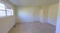 This single-floor residential space presents a straightforward and functional layout designed for comfortable everyday living. The interior is characterized by its clean, neutral color palette, with white walls that create a bright and airy atmosphere throughout. The main living area is generously proportioned, providing ample room for a variety of furniture arrangements to suit different lifestyle needs. Soft, light-toned wall-to-wall carpeting covers the floors, adding warmth and a quiet underfoot feel to the space. The room is equipped with a ceiling-mounted HVAC vent as well as a floor-level return air vent, ensuring efficient climate control year-round. A thermostat is conveniently positioned near the interior door, offering easy access for temperature management. The solid white interior door features standard hardware and is framed cleanly against the surrounding walls. A window provides a source of natural light, contributing to the open and uncluttered feel of the space. The overall ambiance is minimal and move-in ready, offering a blank canvas well-suited for personalization. The simplicity of the design emphasizes practicality, making this a versatile space adaptable to a range of uses and furnishing styles.