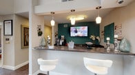 This well-appointed single-floor space blends professional functionality with thoughtful interior design, creating an inviting and visually engaging environment throughout. At the heart of the space is a sleek reception or service counter finished in dark granite, accented by elegant pendant lighting that casts a warm glow. A bold teal-green feature wall behind the counter provides a striking focal point, complemented by a wall-mounted flat-screen display and a curated arrangement of decorative items, including fresh florals and a coffee station. Modern white bar-style seating at the counter adds a clean, contemporary touch.

Beyond the reception area, the space transitions into a more intimate gathering or meeting room centered around a distinctive round green marble table paired with woven rattan chairs. A large, framed abstract artwork in soft neutral tones adorns the wall, adding an artistic sophistication to the room. The floors throughout are finished in a rich, dark polished concrete, lending an industrial-chic character that contrasts beautifully with the warm white walls.

A sliding barn door provides a stylish and functional partition, while a set of antique-style French doors connects to an adjacent area adorned with framed posters and artwork. Recessed ceiling lighting throughout ensures the space is well-illuminated, contributing to the overall warm and welcoming ambiance of this thoughtfully designed interior.