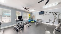 Located at 3401 Bremer Hall Ct in Raleigh, NC, this fitness center is a fully equipped, single-room facility designed to support a wide range of workout routines. The space features soft blue-gray walls, white crown molding, and a durable rubber-floored surface ideal for high-traffic athletic use. Dual ceiling fans and flush fluorescent lighting ensure the room remains well-ventilated and brightly lit throughout. Large windows fitted with white blinds allow natural light to filter in while maintaining privacy. A wall-mounted flat-screen television provides entertainment during workouts, accompanied by a wall clock for time management. The cardio zone includes treadmills, an elliptical trainer, and a stationary spin bike — all TRUE-brand machines. Strength training is supported by a multi-station cable machine, a weight bench, and a tiered dumbbell rack with a comprehensive range of free weights. Functional training accessories are well-represented, with a dedicated rack holding medicine balls, stability balls, resistance bands, and foam rollers. Full-length wall mirrors on one side of the room visually expand the space and assist with exercise form. A fire extinguisher and posted safety guidelines reflect a commitment to user safety. The entrance features double doors, providing convenient access to this comprehensive fitness facility.