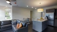 Located at 160 King Ave in the vibrant Short North neighborhood of Columbus, OH, this modern two-floor space offers a thoughtfully designed open-concept layout that maximizes both functionality and style. The main living area showcases dark hardwood-style flooring that flows seamlessly throughout, anchoring the space with a warm yet contemporary feel. Recessed lighting is complemented by a statement drum pendant light above the kitchen island, creating well-distributed illumination throughout. The kitchen features a granite-topped island with gray cabinetry, providing both prep space and a natural gathering point that connects effortlessly to the living and dining areas. The living area is comfortably appointed, while the adjacent dining space accommodates a small round table and chairs, making it ideal for casual meals and entertaining. Neutral wall tones and clean-lined finishes give the space a fresh, move-in-ready aesthetic. Interior doors lead to additional rooms, offering privacy and separation between living spaces. Spanning two floors, the layout provides ample room for a variety of uses. The overall ambiance is clean, contemporary, and practical — well-suited for urban living in one of Columbus's most walkable and dynamic neighborhoods.