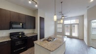 This single-story, one-bedroom, one-bathroom residence in Houston, TX (77025) offers a thoughtfully designed open-concept layout with modern finishes throughout. The kitchen serves as a central highlight, featuring rich dark-espresso cabinetry, granite countertops, a central island with a double-basin sink, and a full suite of black appliances including a range, microwave, and refrigerator with ice maker. Track lighting and pendant lights illuminate the kitchen, while wood-look flooring extends throughout the main living areas, adding warmth and cohesion. The living area benefits from a ceiling fan, recessed lighting, and large French doors that invite natural light and provide access to an outdoor patio. The bedroom is carpeted for comfort and includes oversized windows fitted with blinds, allowing for ample natural light and outdoor views. The bathroom is finished with large-format tile flooring, a granite-topped vanity with dark cabinetry, a well-lit mirror, and a shower-tub combination. Additional storage is provided via a tall pantry cabinet in the kitchen area. The overall design reflects a clean, contemporary aesthetic with a neutral color palette that enhances the sense of openness throughout the unit.