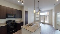 This single-story, one-bedroom, one-bathroom residence in Houston, TX (77025) offers a thoughtfully designed open-concept layout with modern finishes throughout. The kitchen serves as a central highlight, featuring rich dark-espresso cabinetry, granite countertops, a central island with a double-basin sink, and a full suite of black appliances including a range, microwave, and refrigerator with ice maker. Track lighting and pendant lights illuminate the kitchen, while wood-look flooring extends throughout the main living areas, adding warmth and cohesion. The living area benefits from a ceiling fan, recessed lighting, and large French doors that invite natural light and provide access to an outdoor patio. The bedroom is carpeted for comfort and includes oversized windows fitted with blinds, allowing for ample natural light and outdoor views. The bathroom is finished with large-format tile flooring, a granite-topped vanity with dark cabinetry, a well-lit mirror, and a shower-tub combination. Additional storage is provided via a tall pantry cabinet in the kitchen area. The overall design reflects a clean, contemporary aesthetic with a neutral color palette that enhances the sense of openness throughout the unit.