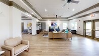 Located at 100 Marcus Byers Dr in Huntsville, Alabama, this thoughtfully designed community space presents a welcoming and polished environment across a single, open floor. The layout flows seamlessly from one area to the next, unified by light hardwood flooring and a consistent palette of soft whites and warm neutrals accented by distinctive dark olive crown molding and tray ceilings throughout.

The expansive living and lounge area serves as the centerpiece of the space, featuring ceiling fans, recessed lighting, and multiple seating arrangements that accommodate both social gatherings and quiet relaxation. A dedicated reading nook with open shelving and accent chairs adds a refined, residential feel. A separate seating alcove provides additional privacy for smaller groups.

The dining area is bathed in natural light from large windows overlooking lush greenery, centering around a round marble-top table that reinforces the space's sophisticated aesthetic. Adjacent to the dining area, a fully equipped kitchen impresses with an expansive quartz island, crisp white shaker cabinetry, stainless steel appliances, and stylish glass pendant lighting — ideal for hosting and communal use.

A functional office area with a desk, computer, and ample natural light rounds out the space, making it well-suited for residents needing a professional work environment. Every area reflects a cohesive, upscale design that balances comfort with practicality.