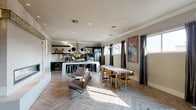 This single-floor space showcases a thoughtfully designed open-concept layout that seamlessly connects a fully equipped kitchen, dining area, and lounge. The herringbone-patterned hardwood flooring flows throughout, lending a sophisticated visual texture that unifies the space.

The kitchen is appointed with dark cabinetry, a mirrored subway tile backsplash, stainless steel appliances, a professional-grade range hood, and a large central island with a white countertop surrounded by clear acrylic bar stools. Recessed lighting and abundant natural light from multiple windows keep the space bright and functional.

The dining area features a substantial wood-top table with gold-accented chairs and a striking multi-arm pendant light overhead. A large colorful artwork anchors the adjacent wall, adding a bold artistic focal point. Dark drapery frames the windows, offering contrast against the neutral grey walls.

The lounge area is furnished with a wraparound gold leather sectional, tufted accent chairs with chrome frames, and sleek gold accent tables. A large pass-through window overlooks what appears to be a building lobby with an elevator, suggesting this space serves a communal or professional purpose. Wall sconces and recessed lighting throughout contribute to a polished, contemporary ambiance.