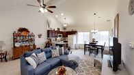 This single-floor home is defined by its dramatic vaulted ceilings that rise steeply above the open-concept main living area, creating an expansive, airy atmosphere throughout. The living room serves as the heart of the space, centered around a comfortable sectional sofa and complemented by a ceiling fan overhead, recessed lighting, and a large area rug that anchors the seating arrangement. The kitchen is positioned along the back wall, featuring rich dark wood cabinetry, white appliances, and a functional layout that integrates seamlessly with the rest of the living space. A pendant light fixture hangs above, adding a warm focal point to the cooking area. Adjacent to the kitchen, the dining area benefits from a large window that floods the space with natural light, with a chandelier overhead and a classic dining table and chair set beneath it. Decorative touches throughout — including seasonal accents, wall-mounted shelving, and carefully placed greenery — give the space a warm, lived-in character. The neutral color palette of soft whites, warm beiges, and muted blues ties each area together cohesively, making the entire floor feel welcoming and well-appointed.