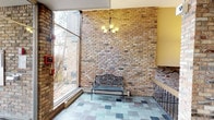 This single-floor space makes a strong first impression through its thoughtfully designed entryway, where classic architectural details and natural light come together to create a warm and inviting atmosphere. The most prominent feature is the expansive exposed brick wall, showcasing warm golden and earthy tones that lend a sense of character and timeless charm to the space. Floor-to-ceiling glass windows flank the entry, flooding the interior with natural light and offering open views of the surrounding outdoor landscape, including mature trees and greenery. The flooring consists of multicolored slate tiles arranged in a varied pattern, adding texture and visual interest underfoot. A wrought iron chandelier with warm-toned bulbs hangs as a central decorative element, complementing the rustic aesthetic of the brick. A decorative wrought iron bench is positioned along the brick wall, providing both a functional and ornamental touch. Potted greenery near the windows softens the industrial feel of the brick and adds a lively natural element to the interior. The glass entry doors open to a tiled vestibule, reinforcing the building's accessible and welcoming design. Overall, the space balances rustic and refined elements, creating an entryway that is both functional and aesthetically distinctive.