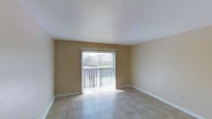 This single-floor space presents a clean, functional living area finished with light-toned ceramic tile flooring throughout. The room is characterized by its simplicity and openness, with neutral beige walls and white baseboards that create a bright, airy atmosphere. A textured popcorn ceiling adds a subtle layer of detail overhead.

The most prominent feature of the space is a full-width sliding glass door fitted with vertical blinds, which serves as the primary source of natural light. The door opens to an outdoor area bordered by a fence, offering a degree of privacy while maintaining a connection to the exterior. Sunlight filters through the blinds, casting linear patterns across the tile floor and contributing to the room's welcoming feel.

The walls are kept simple with minimal fixtures, and the layout is open and versatile, lending itself well to a variety of furniture arrangements. The overall ambiance is modest and practical, emphasizing cleanliness and functionality. The combination of natural light, neutral tones, and durable tile flooring makes the space easy to maintain and adaptable to different interior styles.