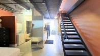 This two-story loft showcases a distinctive industrial aesthetic throughout its open-concept layout. The exposed ceiling reveals structural ductwork and corrugated metal detailing, lending an authentic urban character to the space. Polished concrete floors run continuously across the main level, reinforcing the raw, warehouse-inspired design.

A striking open-riser metal staircase with a dark finish commands attention along one wall, providing access to the upper floor while serving as a bold architectural feature. An accent wall in a warm orange-red hue adds a pop of color that contrasts effectively with the otherwise neutral industrial palette.

The main level integrates multiple functional areas in close proximity. A kitchen with dark cabinetry and stainless steel appliances is visible along one corridor, leading into a living area furnished with a sofa and ambient lighting. A sleeping area is tucked to one side, accompanied by a full bathroom enclosed with glass partition elements, maintaining an open yet defined sense of separation.

Track lighting mounted to the ceiling provides practical illumination throughout, enhancing the loft's modern, urban character.