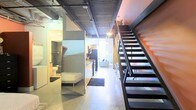 This two-story loft showcases a distinctive industrial aesthetic throughout its open-concept layout. The exposed ceiling reveals structural ductwork and corrugated metal detailing, lending an authentic urban character to the space. Polished concrete floors run continuously across the main level, reinforcing the raw, warehouse-inspired design.

A striking open-riser metal staircase with a dark finish commands attention along one wall, providing access to the upper floor while serving as a bold architectural feature. An accent wall in a warm orange-red hue adds a pop of color that contrasts effectively with the otherwise neutral industrial palette.

The main level integrates multiple functional areas in close proximity. A kitchen with dark cabinetry and stainless steel appliances is visible along one corridor, leading into a living area furnished with a sofa and ambient lighting. A sleeping area is tucked to one side, accompanied by a full bathroom enclosed with glass partition elements, maintaining an open yet defined sense of separation.

Track lighting mounted to the ceiling provides practical illumination throughout, enhancing the loft's modern, urban character.