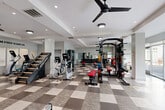 Located at 9142 Richmond Hwy in Fort Belvoir, VA, this fitness center offers a generously sized, single-floor exercise room designed to support a comprehensive workout experience. The space features a striking checkerboard-patterned tile floor in contrasting shades of gray and beige, lending a bold, modern aesthetic throughout. Recessed lighting and large ceiling fans maintain a bright, comfortable environment, while expansive floor-to-ceiling windows along one wall flood the room with natural light and provide views of the surrounding greenery outside.

The exercise room is fully equipped for both cardio and strength training. Cardio offerings include treadmills, ellipticals, stationary bikes, a rowing machine, and a stair climber. The strength training area features a multi-functional cable machine, a power rack loaded with a full set of weight plates, adjustable benches, and a dedicated dumbbell rack with wall-mounted mirrors for form feedback. Motivational wall decals, including "Live Here, Train Here" and "No Stress to Enjoy Life," enhance the energizing atmosphere. A wall-mounted flat-screen TV adds to the amenity appeal. The clean white walls, coffered ceiling details, and structured layout make this fitness center both functional and visually polished.