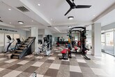 Located at 9142 Richmond Hwy in Fort Belvoir, VA, this fitness center offers a generously sized, single-floor exercise room designed to support a comprehensive workout experience. The space features a striking checkerboard-patterned tile floor in contrasting shades of gray and beige, lending a bold, modern aesthetic throughout. Recessed lighting and large ceiling fans maintain a bright, comfortable environment, while expansive floor-to-ceiling windows along one wall flood the room with natural light and provide views of the surrounding greenery outside.

The exercise room is fully equipped for both cardio and strength training. Cardio offerings include treadmills, ellipticals, stationary bikes, a rowing machine, and a stair climber. The strength training area features a multi-functional cable machine, a power rack loaded with a full set of weight plates, adjustable benches, and a dedicated dumbbell rack with wall-mounted mirrors for form feedback. Motivational wall decals, including "Live Here, Train Here" and "No Stress to Enjoy Life," enhance the energizing atmosphere. A wall-mounted flat-screen TV adds to the amenity appeal. The clean white walls, coffered ceiling details, and structured layout make this fitness center both functional and visually polished.