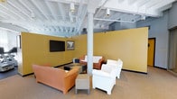This single-floor space offers a welcoming lounge environment designed for comfortable gathering and relaxation. The room is defined by bold mustard-yellow walls that create a warm, energetic atmosphere, contrasted by a white-painted exposed ceiling that reveals structural beams, ductwork, and track lighting. The industrial-style ceiling treatment adds visual interest while contributing to a sense of openness and height overhead.

The space is furnished with a thoughtfully arranged mix of seating, including a terracotta-toned sofa, white armchairs, and a rust-colored chair grouping, offering flexible configurations for both small conversations and larger gatherings. A patterned, checkered ottoman set serves as a central coffee table, adding a playful design element to the arrangement.

A wall-mounted flat-screen television is positioned on one of the yellow accent walls alongside framed artwork, making the space suitable for presentations or media viewing. A structural column anchors the center of the room, subtly dividing seating zones. Neutral carpeted flooring ties the design together, and directional track lighting ensures the space is well-illuminated throughout.