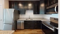 Located at 3730 Evergreen Dr in Monroeville, PA, this single-floor residence offers a thoughtfully designed layout with contemporary finishes throughout. The kitchen is a standout feature, showcasing rich dark cabinetry, light granite countertops, and a full suite of stainless steel appliances including a refrigerator, electric range with over-the-range microwave, and dishwasher. A kitchen island adds additional counter space and functionality. The open living area is bathed in natural light through large windows and a glass door that leads to a private outdoor balcony, offering pleasant views of surrounding greenery. Warm hardwood-style flooring runs throughout the main living spaces, lending a cohesive and modern feel. The bedrooms are carpeted for comfort, with one featuring a walk-in closet with built-in shelving and direct access to a full bathroom. A second bedroom benefits from a large window with natural light. Two bathrooms serve the unit, both featuring classic black-and-white mosaic tile flooring and dark vanity cabinetry. One bathroom includes a walk-in shower, while the other offers a single-sink vanity. The overall aesthetic is clean, modern, and move-in ready, with crisp white walls and consistent finishes throughout.