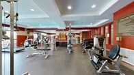 This fitness center is organized across two dedicated exercise rooms, offering a comprehensive workout environment for users of all fitness levels. The main exercise room is a generously sized space characterized by bold red accent walls, a coffered ceiling with recessed lighting, and durable rubber flooring. Full-length mirrors line one wall, amplifying the sense of space and allowing for form checks during workouts. The room is outfitted with a wide array of strength training equipment, including multiple Hoist-brand machines, a cable station, a dumbbell rack, and adjustable benches. Large windows along one side admit abundant natural light, offering views of the surrounding exterior. Decorative diamond-shaped wall art adds a stylish design element to the space. The secondary exercise room features warm hardwood flooring and a more intimate setting, housing a Peloton spin bike and additional strength equipment. This room connects to adjacent common areas through glass partitions, maintaining an open, airy feel. Cardio equipment, including Matrix treadmills and a stationary bike, is positioned near expansive windows to provide an engaging outdoor view during workouts. Together, the two rooms form a cohesive, well-appointed fitness facility designed for both functionality and visual appeal.