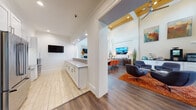 This single-floor community space is designed with an open, airy layout and impressive double-height ceilings that create a grand sense of scale throughout. The main lounge area is anchored by a dramatic floor-to-ceiling stacked stone fireplace with a wall-mounted television, flanked by tall windows dressed with white blinds that flood the room with natural light. Comfortable seating, including teal sofas and modern swivel chairs atop vibrant accent rugs, gives the space a welcoming, contemporary feel. Bold abstract artwork adorns the walls, adding color and visual interest.

Adjacent to the lounge, a casual dining area features a rustic wood table with teal upholstered chairs, positioned near an open kitchen bar. The kitchen includes white cabinetry with under-cabinet lighting, a mosaic tile backsplash, and a long counter with stylish bar stools — ideal for casual gatherings. A dedicated coffee and refreshment station with built-in appliances provides added convenience.

A secondary hallway area features white cabinetry with a countertop workspace and a wall-mounted display, serving as a functional support zone. Rich dark hardwood flooring runs throughout and contrasts beautifully with the warm neutral wall tones. The overall ambiance is polished, modern, and inviting — well-suited for community gatherings and social interaction.