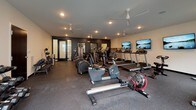 Located at 6847 Stewart Rd in Cincinnati, Ohio, this fitness center offers a well-appointed, single-floor exercise facility designed to support a full range of workout routines. The space is outfitted with durable rubber flooring throughout, providing a safe and comfortable surface for exercise.

The cardio section is equipped with multiple treadmills, each paired with a wall-mounted flat-screen television for entertainment during workouts. Additional cardio options include elliptical trainers, a rowing machine, and stationary bikes, providing a variety of aerobic exercise choices. Ceiling fans are installed overhead to help maintain a comfortable environment during high-intensity training.

A dedicated strength training area features a multi-station cable machine, a full dumbbell rack with a wide range of weights, and kettlebells, catering to both beginners and experienced lifters. The space also includes a bench for additional exercise versatility.

Amenities include a water fountain and a neatly stocked towel station with cubbies, ensuring convenience for users. Glass double doors lead to an outdoor area, allowing natural light to fill the space and offering views of an adjacent outdoor patio. Dark accent walls add a modern, focused aesthetic to the facility. A separate room with glass-paneled doors provides additional enclosed space for specialized equipment or private training.