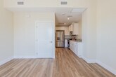 This single-floor unit offers a clean, modern design defined by warm hardwood-style flooring, crisp white trim, and soft neutral walls that create a bright and welcoming atmosphere throughout. The open living area is generously proportioned, bathed in natural light from a large window with blinds that frames a lush outdoor view. A flush ceiling light fixture adds a polished touch to the space.

The kitchen flows seamlessly from the living area and is thoughtfully designed with white shaker-style cabinetry extending to the ceiling, smooth light-toned countertops, and a full suite of stainless steel appliances, including a gas range, over-the-range microwave, refrigerator, and dishwasher. Recessed lighting illuminates the workspace, and a single-basin sink completes the functional layout.

The bathroom continues the home's clean aesthetic, featuring light wood-style flooring, a white vanity with an integrated sink, and a large mirror. A window fitted with slatted blinds allows natural light to filter in while maintaining privacy. A tankless water heater is neatly integrated into the space.

The entry area provides a welcoming threshold with access to an adjacent outdoor space, reinforcing the unit's bright, airy feel. Overall, the space presents a cohesive, contemporary design with quality finishes and a practical, comfortable layout.