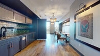 This thoughtfully designed single-floor social space combines upscale aesthetics with functional entertainment areas. Warm hardwood flooring flows throughout, unifying the distinct zones within the open layout. A striking wet bar anchors one side of the room, featuring dark cabinetry with mixed metal hardware, stainless steel countertops, and a bold iridescent teal hexagonal tile backsplash that serves as a dramatic focal point. Mesh-front upper cabinets and open shelving complete the bar's sophisticated design. Adjacent to the bar, a dedicated game area showcases a round poker-style table with built-in cup holders, surrounded by upholstered chairs beneath an eye-catching fringe chandelier. Floor-to-ceiling windows flood this corner with natural light and offer views of the surrounding neighborhood. The space also features a cozy lounge nook with wingback chairs and a hammered copper accent table set against patterned geometric wallcovering. A reception-style counter with teal cabinetry and mirrored upper panels defines another distinct area, flanked by plush barrel chairs. Throughout the space, layered lighting — including pendant chandeliers and recessed fixtures — creates an inviting ambiance suited for both casual gatherings and organized social events.
