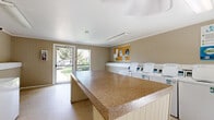 This shared laundry facility is a thoughtfully organized, single-floor space designed to accommodate all essential laundry needs efficiently. The room features a row of coin-operated top-loading washing machines along one wall and a bank of stacked coin-operated dryers along another, providing ample capacity for multiple users. A prominent, generously sized folding counter with a granite-style surface runs centrally through the space, offering a convenient area for sorting and folding laundry. The floors are finished with light-toned tile, contributing to a clean and practical atmosphere throughout. Natural light enters through several windows, including views of surrounding greenery, brightening the space and creating an open, welcoming feel. A cork bulletin board is mounted near the entrance, providing a space for community notices and information. A beverage vending machine is conveniently situated within the room, adding a practical amenity for users. Overhead fluorescent lighting ensures the space remains well-lit at all hours. The walls feature a two-tone treatment with wainscoting, maintaining a tidy and organized appearance. A glass-panel entry door allows additional natural light and easy outdoor visibility. Overall, the facility is clean, functional, and user-friendly.