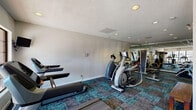 This fitness center is a single-floor exercise facility designed to support a well-rounded workout routine. The space is outfitted with a variety of cardio equipment, including treadmills, an elliptical trainer, and a stationary bike, all arranged to allow comfortable circulation throughout the room. A multi-station cable strength machine anchors one section of the facility, accompanied by a free weight area with dumbbells and a dedicated weight bench, offering options for both resistance and functional training. Full-length mirrors line a portion of the walls, visually expanding the space and allowing users to monitor their form. The flooring features a distinctive teal and charcoal patterned carpet tile, adding a modern aesthetic contrast to the neutral gray walls. Recessed lighting and a textured ceiling provide a clean, functional overhead finish. Multiple windows along one wall allow natural light to enter and offer views of the surrounding outdoor area, while wall-mounted televisions provide entertainment during workouts. A restroom is accessible directly from the facility, adding convenience for users. The overall layout is practical and thoughtfully organized, providing a comprehensive fitness environment.