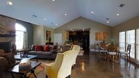 This single-story space makes a striking impression with its dramatic vaulted ceilings that rise to a sharp peak, creating an expansive sense of volume throughout the main living area. Recessed lighting is thoughtfully distributed across the ceiling, providing warm, even illumination that complements the open-concept layout. The polished concrete floors lend an industrial-chic aesthetic while offering a durable, low-maintenance surface that flows continuously through the space. The living area is anchored by a plush gray sofa accented with rich red throw pillows, paired with distinctive woven accent chairs in a warm golden hue, all arranged around a sleek coffee table atop a layered area rug. Wall art adds curated personality to the neutral, earthy-toned walls. Transitioning naturally from the living area, a dedicated dining space features a solid wood table surrounded by matching chairs, offering a comfortable setting for gatherings. Beyond, the kitchen is set within a more intimate, lower-ceilinged alcove, outfitted with dark cabinetry, stainless appliances, and under-cabinet lighting that creates a cozy contrast to the airy main space. The overall ambiance is warm, modern, and inviting, with a cohesive design that balances comfort and style throughout.
