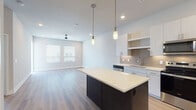 This single-floor residence showcases a thoughtfully designed open-concept layout with a clean, contemporary aesthetic. The kitchen serves as the focal point, anchored by a substantial center island with a dark wood base and bright white countertop, illuminated by sleek cylindrical pendant lights. White flat-front cabinetry extends to the ceiling, complemented by a neutral tile backsplash and a full suite of stainless steel appliances, including a French door refrigerator, electric range, over-the-range microwave, and dishwasher. Light wood-look flooring runs throughout the kitchen and living areas, lending warmth to the bright, neutral color palette. The living area is generously proportioned, featuring a ceiling fan with recessed lighting, and access to an outdoor balcony. Bedrooms are finished with plush carpeting and are served by a well-appointed bathroom featuring a single-sink vanity with white countertops, a framed mirror, wood-look flooring, and a glass-enclosed shower. Accessibility-conscious grab bars are present in the bathroom. A dedicated entry foyer with a paneled door provides a welcoming transition into the space.