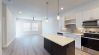 This single-floor residence showcases a thoughtfully designed open-concept layout with a clean, contemporary aesthetic. The kitchen serves as the focal point, anchored by a substantial center island with a dark wood base and bright white countertop, illuminated by sleek cylindrical pendant lights. White flat-front cabinetry extends to the ceiling, complemented by a neutral tile backsplash and a full suite of stainless steel appliances, including a French door refrigerator, electric range, over-the-range microwave, and dishwasher. Light wood-look flooring runs throughout the kitchen and living areas, lending warmth to the bright, neutral color palette. The living area is generously proportioned, featuring a ceiling fan with recessed lighting, and access to an outdoor balcony. Bedrooms are finished with plush carpeting and are served by a well-appointed bathroom featuring a single-sink vanity with white countertops, a framed mirror, wood-look flooring, and a glass-enclosed shower. Accessibility-conscious grab bars are present in the bathroom. A dedicated entry foyer with a paneled door provides a welcoming transition into the space.