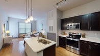 This single-floor unit offers a thoughtfully arranged layout that maximizes its footprint. The open-concept kitchen serves as a central highlight, featuring a large island with a double stainless steel sink, pendant lighting overhead, and dark espresso cabinetry that contrasts cleanly against white countertops. Stainless steel appliances — including a range, microwave, and refrigerator — complete the kitchen. Light hardwood flooring flows throughout the main living area, and track lighting adds a modern touch to the soaring ceiling height visible in the kitchen and living space. The living area, visible from the kitchen, benefits from natural light streaming through large windows fitted with horizontal blinds.

The bedroom is carpeted and comfortably sized, featuring multiple windows with sheer draping curtains that allow natural light while maintaining privacy. A ceiling fan adds functionality to the space. The full bathroom includes a subway-tiled tub and shower combination, a dark-cabinetry vanity with a white countertop, and large-format tile flooring. A hallway with closet access connects the bedroom and bathroom, contributing to an efficient and practical flow throughout the unit.