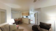 This single-floor residence is designed with an open-concept layout that seamlessly integrates the living, dining, and kitchen areas into one cohesive space. The living area is carpeted and furnished with comfortable seating, while a ceiling fan overhead provides ventilation and contributes to the relaxed atmosphere of the room. Adjacent to the living area, a dining space accommodates a wooden table and chair set, making it well-suited for everyday meals or casual gatherings. A small floral arrangement on the table adds a touch of warmth to the area.

The kitchen features a raised peninsula counter that separates it from the dining area while maintaining an open sightline throughout the space. Light wood cabinetry provides storage above, and basic appliances are visible along the counter. The neutral color palette of white walls and beige carpet runs consistently throughout, contributing to a clean, understated aesthetic. A hallway visible from the main living area leads to additional rooms, with a door positioned at the end suggesting access to a private area or entry point. Blinds on the windows offer light control and privacy.